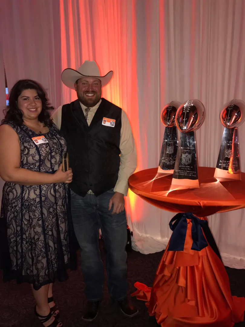 Brian with his wife April at the 50th anniversary party for the Denver Broncos Cheerleaders standing next to the Denver Broncos 3 Lombardi trophies